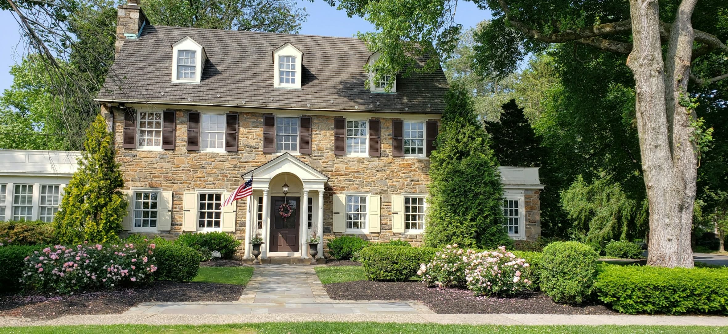 A stone-and-shingle Dutch Colonial home with a white-columned entry portico, dormer windows, and mature flowering landscaping in warm afternoon light